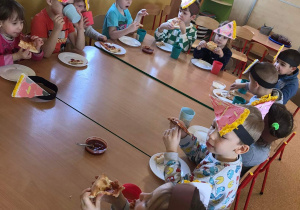Dzieci siedzą przy długim stole i jedzą pizzę.