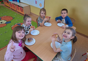 Dzieci siedzą przy stoliku i jedzą pączki.