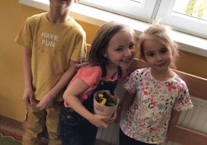 Chłopiec i dwie dziewczynki pozują do zdjęcia - dziewczynka trzyma doniczkę z posadzonym bratkiem.