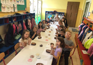 Grupa dzieci siedzi przy długim stole i dekoruje czekoladowe jajeczka.