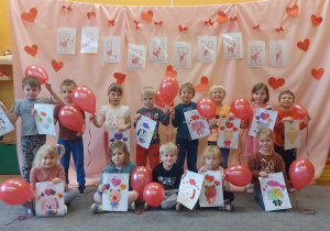 Dzieci trzymają w ręce czerwone balony w kształcie serca i pozują razem do zdjęcia na różowym tle