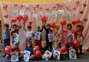 Dzieci trzymają w ręce czerwone balony w kształcie serca i pozują razem do zdjęcia