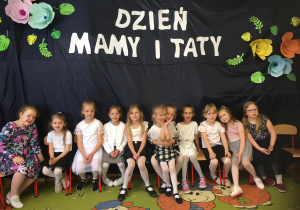 Dziewczynki siedzą pod napisem "Dzień Mamy i Taty".