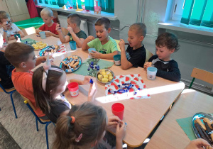 Dzieci jedzą poczęstunek przy stolikach.