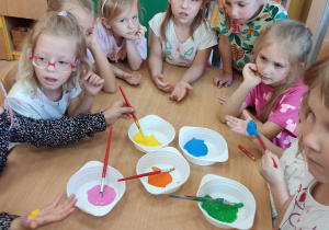 Dzieci siedzą przy stoliku, na którym są ustawione farby.