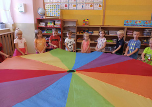 Dzieci stoją w kole trzymając w rękach kolorową chustę animacyjną
