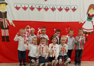 Dzieci ubrane w stroje galowe trzymają w ręce kokardy narodowe i pozują w grupie do zdjęcia na tle biało - czerwonego materiału z napisem 11 listopada