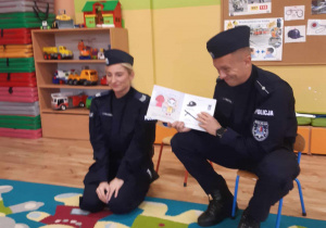 Pan policjant pokazuje dzieciom książeczkę, obok niego siedzi pani policjantka.
