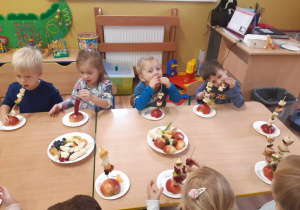 Dzieci siedzą przy stolikach i zjadają owocowe koreczki.