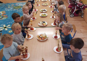 Dzieci siedzą przy stolikach, przed sobą maja talerzyki a na nich owoce nadziane na wykałaczki.