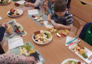 Dzieci siedzą przy stolikach i kroją nożykami obrane owoce.