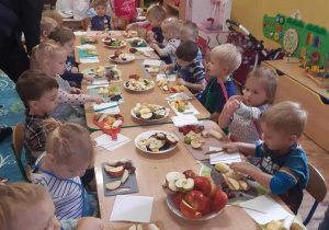 Dzieci siedzą przy stolikach, przed sobą maja różne owoce oraz deseczki do krojenia i nożyki.