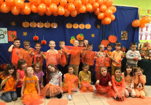Grupa dzieci ubranych w pomarańczowe stroje pozuje do zdjęcia na tle tematycznej - dyniowej ścianki.