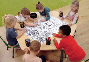 Dzieci siedzą przy stolikach i kolorują sylwetę misia