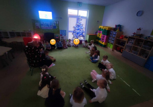 Dzieci siedzą w kole, a nauczyciel podświetla obrazki prezentów