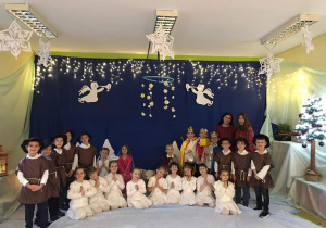 Grupa dzieci w strojach aniołów i pasterzy, wraz z Maryją i Józefem oraz Trzema Królami pozują na tle dekoracji przygotowanej na Jasełka.