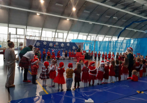 Dzieci ubrane na czerwono w mikołajkowych czapkach stoją w dużym kole.