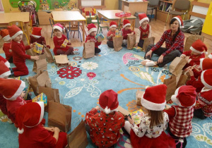 Dzieci ubrane na czerwono w mikołajkowych czapkach stoją w dużym kole, trzymają przed sobą torby z prezentami .