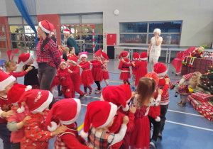 Dzieci ubrane na czerwono w mikołajkowych czapkach tańczą, tworząc sznur wagoników