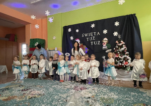 Dzieci wraz z paniami przebrane za aniołki pozują do zdjęcia. W tle dekoracja świąteczna z napisem, Święta tuż, tuż..."