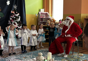 Dzieci przebrane za aniołki śpiewają dla Mikołaja, który siedzi przed nimi na krześle.