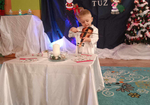 Chłopiec ubrany na biało stoi przed wigilijnym stołem i gra na skrzypcach. W tle świąteczna dekoracja