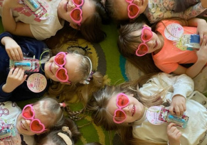 Dziewczynki w różowych okularach pozują do zdjęcia leżąc na dywanie.