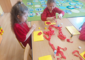 Dwie dziewczynki wyklejają serce czerwoną bibułą.