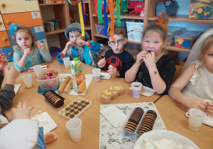 Dzieci przebrane w stroje karnawałowe siedzą przy stoliku i się częstują słodkościami.