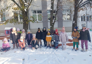 Grupa dzieci ubrana w zimowe ubrania pozuje do zdjęcia w przedszkolnym ogródku.
