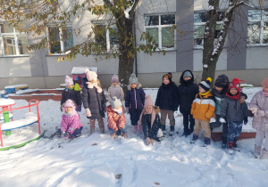 Grupa dzieci ubrana w zimowe ubrania pozuje do zdjęcia w przedszkolnym ogródku.