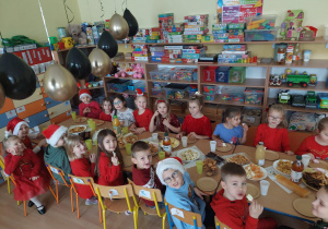 Dzieci w strojach i czapkach Mikołajów siedzą przy stolikach i jedzą słodki poczęstunek.