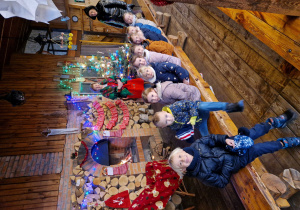 Dzieci siedzą na drewnianej, długiej ławce, przed murowanym kominkiem, w którym pali się drewno