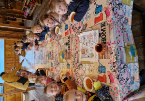 Dzieci siedzą przy długim, drewnianym stole i jedzą zupę pomidorową