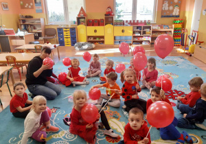 Dzieci siedzą na dywanie, trzymają w rękach czerwone balony. Pani nadmuchuje balony dla dzieci siedząc z nimi w kole.