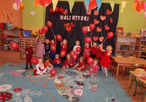 Dzieci ubrane na czerwono z balonami w kształcie serc pozują do zdjęcia. W tle napis ,,WALENTYNKI"