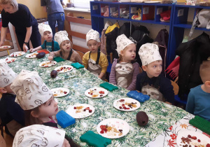 Dzieci ubrane w kucharskie czapki siedzą przy stolikach. Przed nimi ustawione są talerzyki z owocami, chrupkami i wiórkami kokosowymi