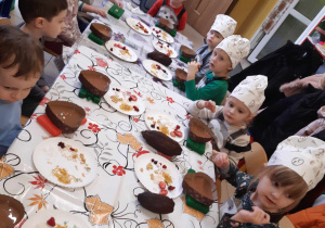 Dzieci ubrane w kucharskie czapki siedzą przy stolikach. Przed nimi ustawione są foremki z czekoladą do dekoracji