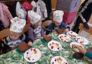 Dzieci ubrane w kucharskie czapki siedzą przy stolikach i dekorują foremki z czekoladą