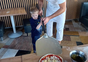 Chłopiec wraz z panem kucharzem wkłada pizzę na szuflę