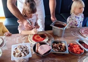 Dziewczynka wraz z kucharzem nakłada sos pomidorowy na pizzę.