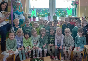 Dzieci pozuja do zdjęcia, pani trzyma kukłę ze słomy, przed dziećmi stoją w pudle kwiaty