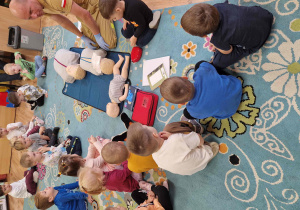 Dzieci siedzą na dywanie, pośrodku leży fantom, na którym dzieci uczą się wykonywać rko