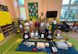 Dzieci siedzą w grupie w nauczycielkami i strażakiem, przed nimi leżą fantomy do ćwiczeń resustytacji