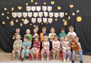 Dzieci siedzą w grupie, trzymają swoje pluszowe misie. W tle napis Dzień Pluszowego Misia na zawieszony na czarnej tkaninie