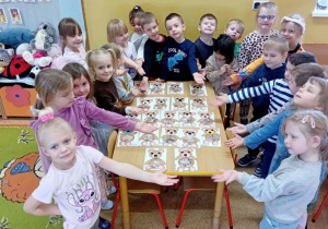 Dzieci siedzą wokół stolika na którym leżą pracę plastyczne przedstawiające pluszowego misia