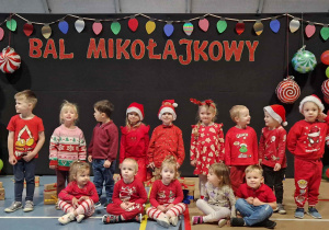 Grupa dzieci w czerwonych strojach pozuje do zdjęcia. W tle napis Bal Mikołajkowy