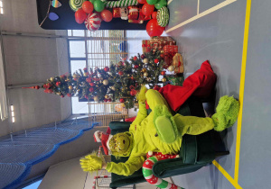 Grinch siedzi na zielonym fotelu, w tle choinka