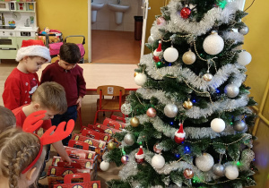 Dzieci stoją przed choinką i oglądają prezenty.