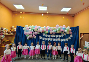 Dzieci stoją w półkolu, dziewczynki mają białe bluzki i rajstopy oraz różowe spódnice, chłopcy mają czarne spodnie i białe koszule. W tle napis pasowanie na przedszkolaka na granatowym tle